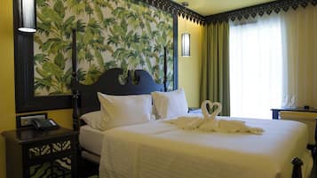 Una habitación de hotel con cama, mesita de noche, papel tapiz tropical en la pared y ventana con cortinas.