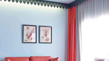 Habitación de hotel con un sofá de cuero rojo, dos láminas botánicas enmarcadas en la pared, un jarrón con flores y una cama con ropa blanca.