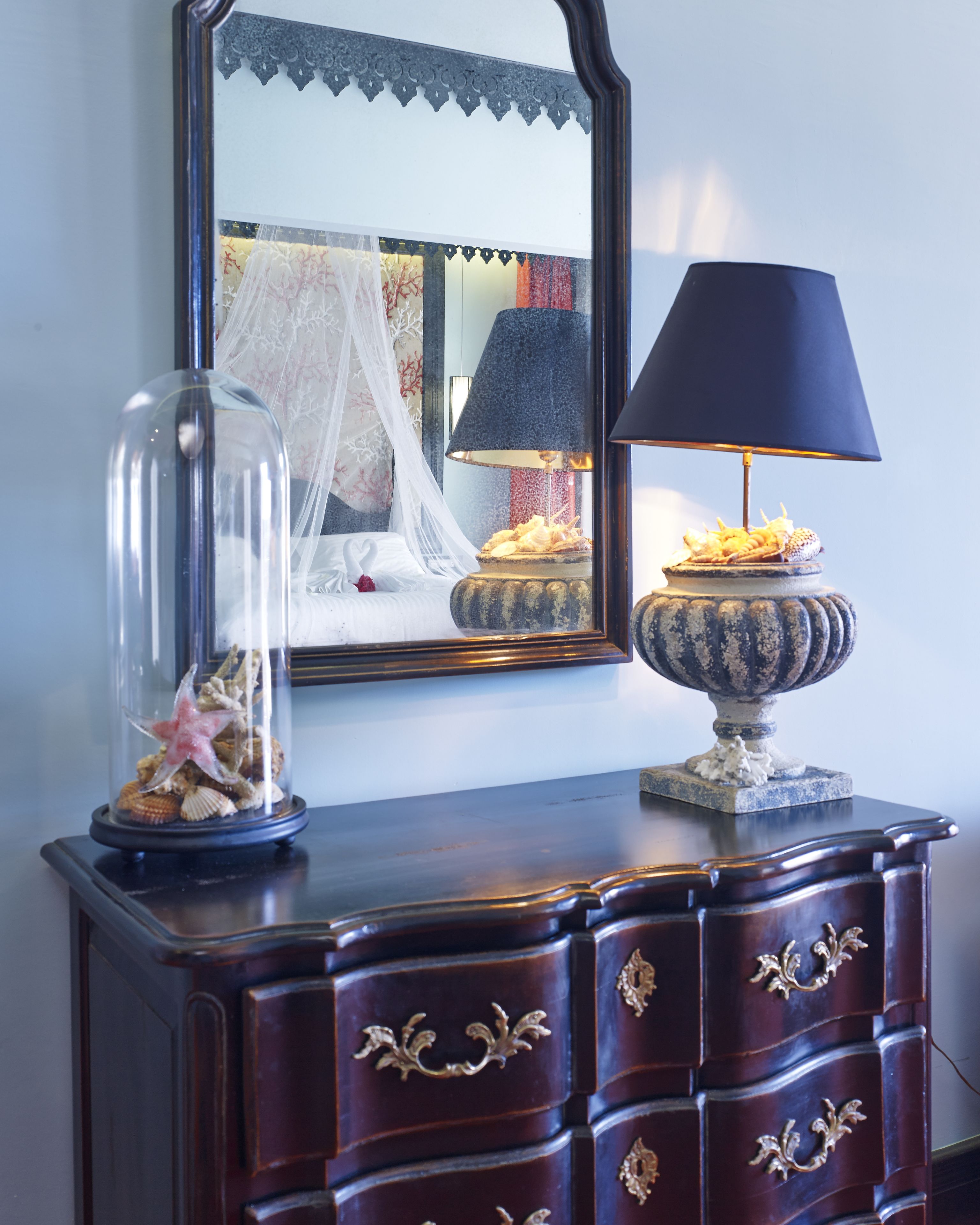 A wooden dresser with ornate gold handles, a glass dome containing seashells and a starfish, and a table lamp with a blue shade.