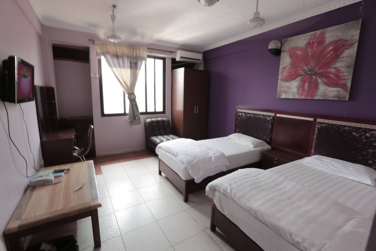 A hotel room with two beds, a desk, a wardrobe, and a large window with curtains.