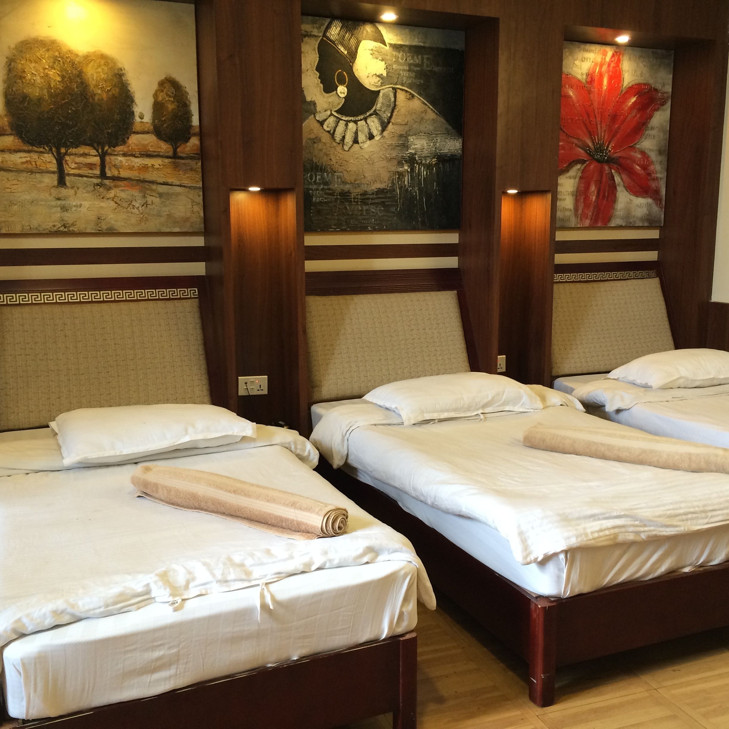 Une chambre d’hôtel avec trois lits parfaitement faits, chacun agrémenté de draps et de serviettes blancs. La chambre est dotée de têtes de lit en bois et d’un grand tableau représentant une fleur rouge accroché au mur.