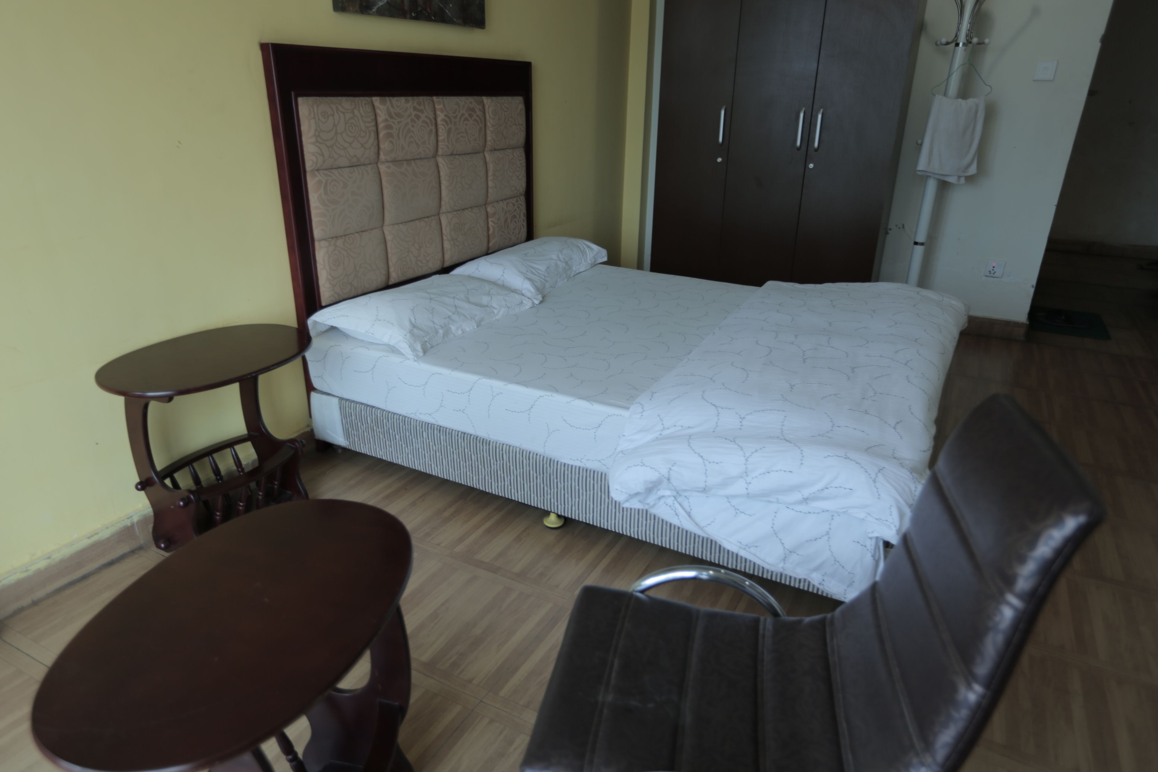 Una cama bien hecha con cabecera, una mesita redonda pequeña y un banco.
