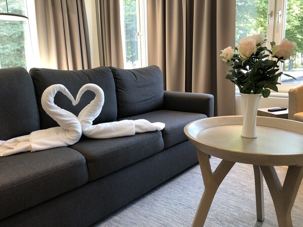 Un sofá gris oscuro con una toalla con forma de corazón y un jarrón blanco con flores sobre una mesita redonda.