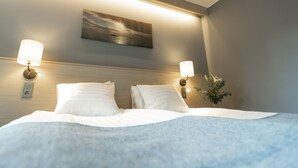 Una cama bien hecha con sábanas blancas, dos lámparas fijadas a la pared, un cuadro sobre la cama y una planta pequeña en la mesita de noche.
