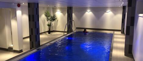 Una piscina interior con un elemento acuático iluminado en azul, una escalera y una planta en maceta.