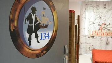 A room with a circular wall decoration featuring a pirate and a ship, a wooden door, a bed with an orange blanket, and a wall with a map and pirate-themed decor.