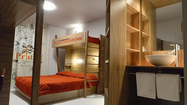 Una cama individual con colchón rojo, cabecera de madera y una pared con un cartel. Hay un estante de madera con un lavabo y un espejo encima.