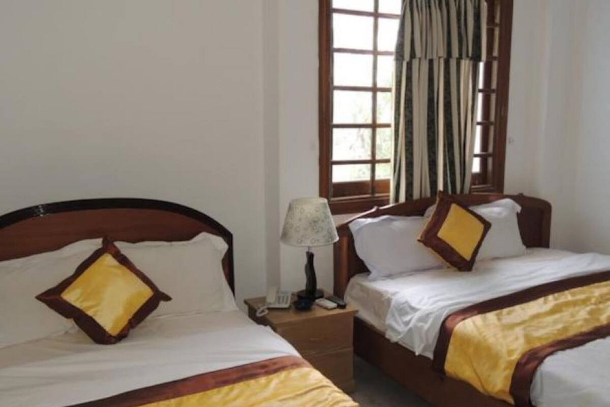 Habitación de hotel con dos camas, cada una con cabecero de madera y ropa de cama blanca. Cuenta con una ventana con cortinas, una mesita de noche con lámpara y un teléfono.