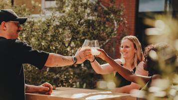 Two people toasting with glasses at an outdoor wooden table with a brick wall and greenery in the background.