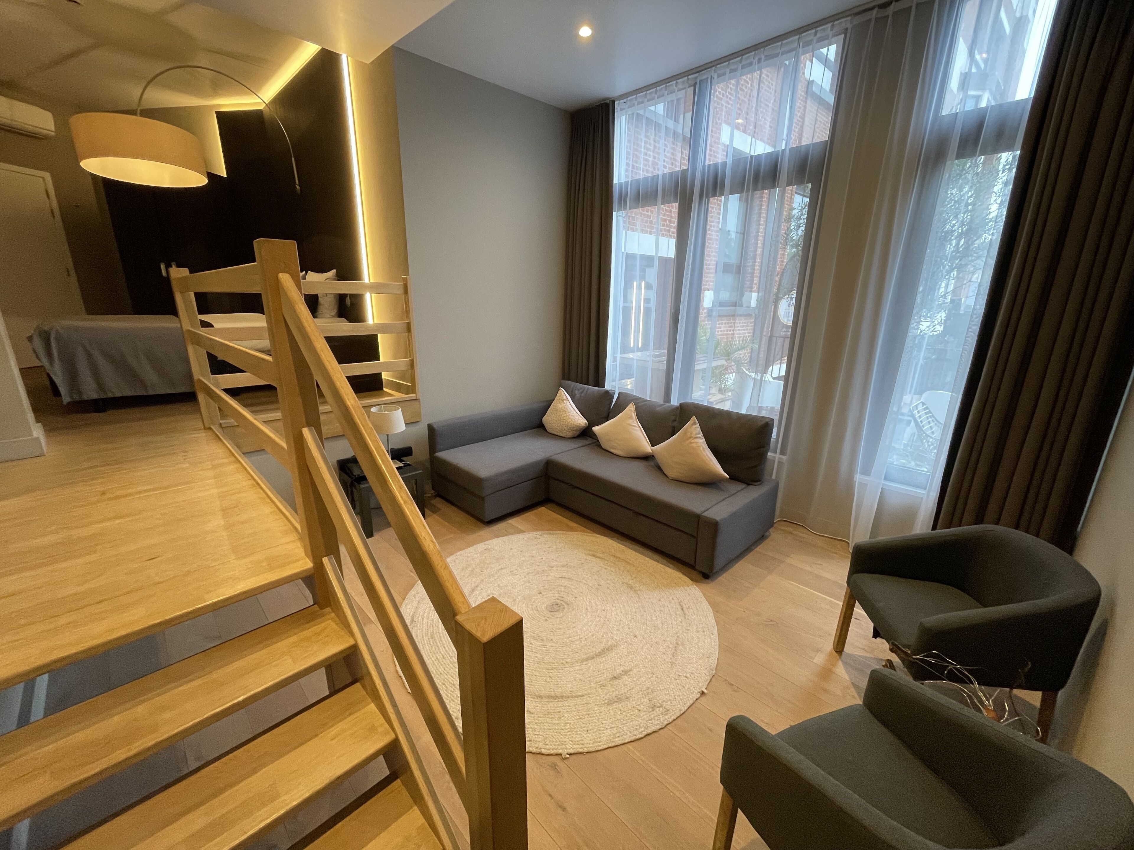 A modern living room with a wooden staircase, a grey sofa with cushions, a black armchair, and large windows with sheer curtains.