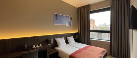 A hotel room with a bed, bedside table, lamp, and a view of the city through a window.