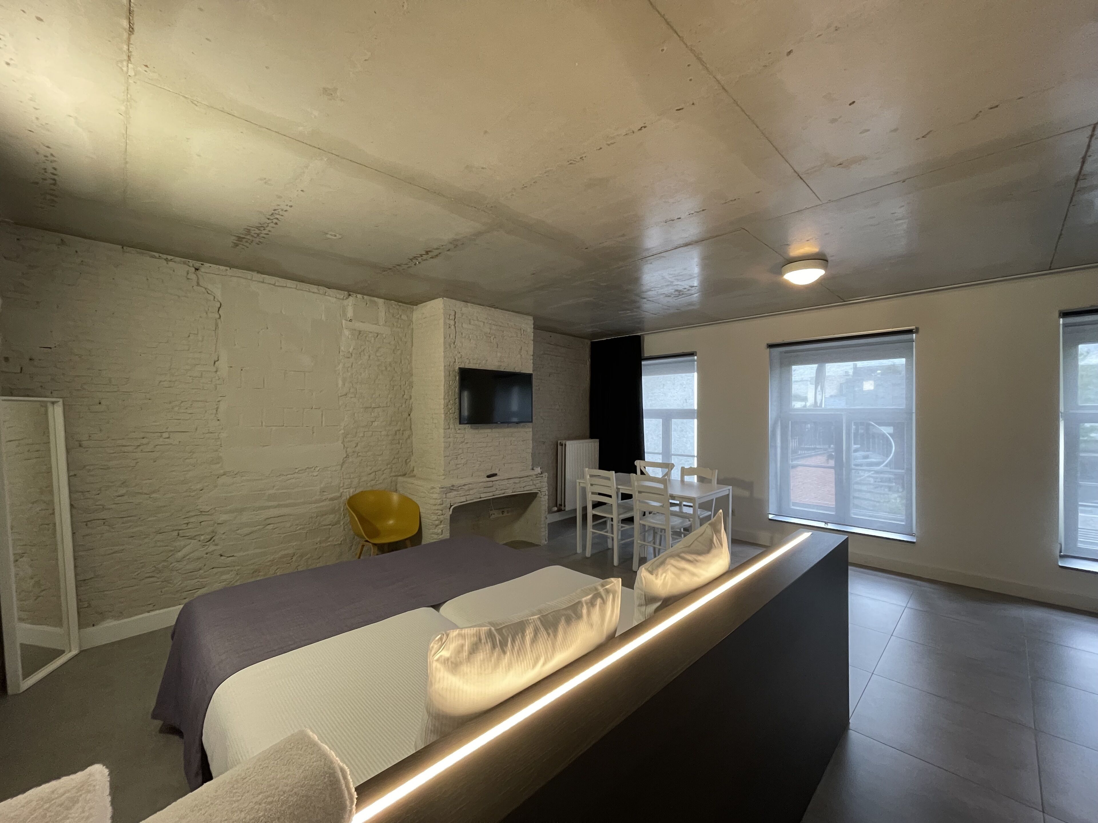 A modern bedroom with a bed, a TV mounted on the wall, and large windows.