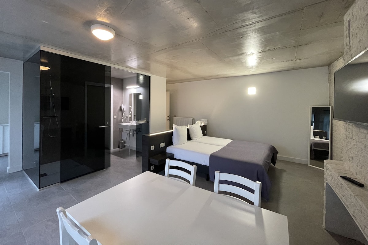 Ein modernes Hotelzimmer mit einem Bett, Nachttischen, einem Schreibtisch und einem Stuhl. Es gibt ein Badezimmer mit Spiegel und Waschbecken.