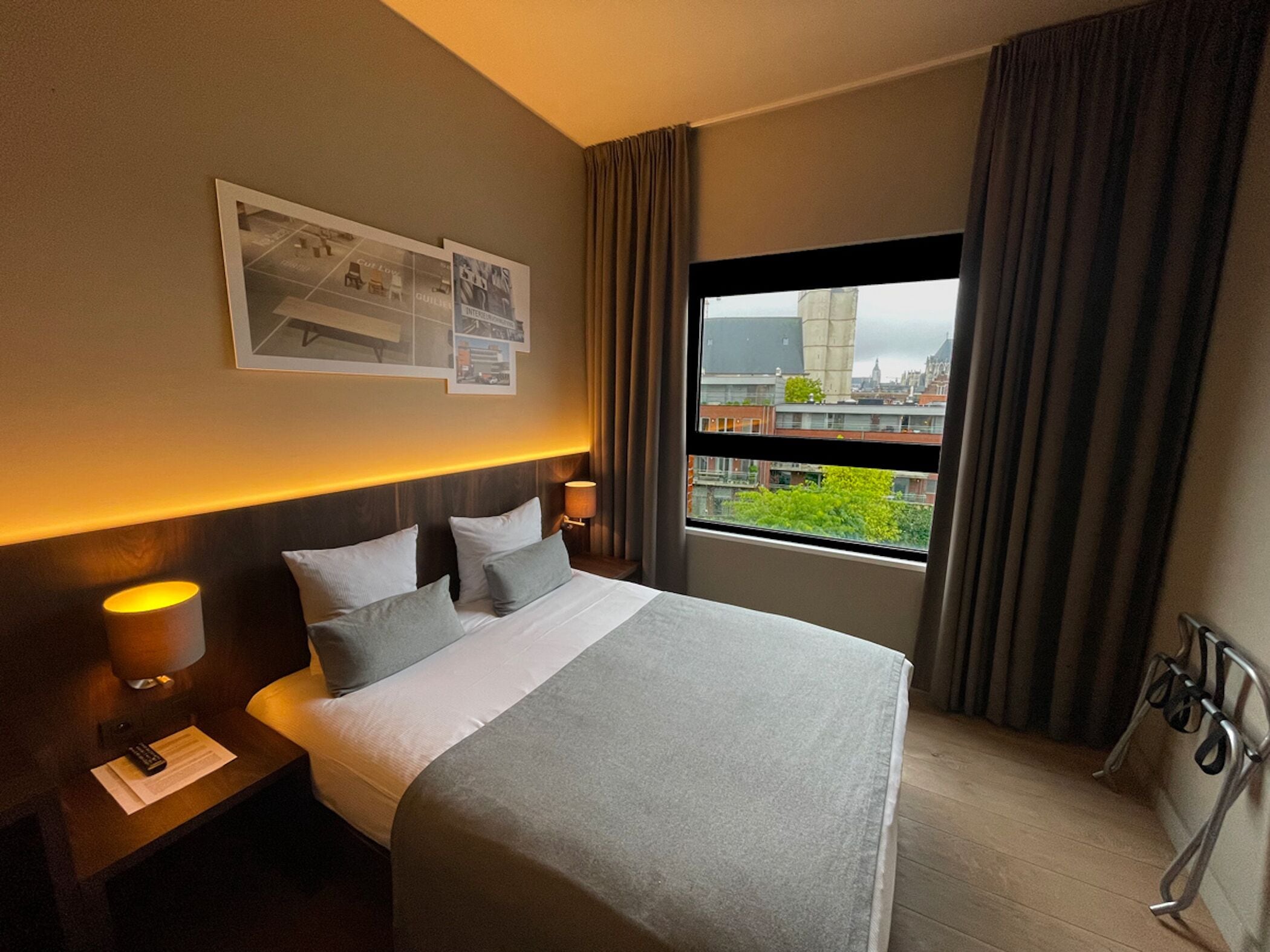 A hotel room with a bed, nightstands, a lamp, a window with curtains, and a city view.