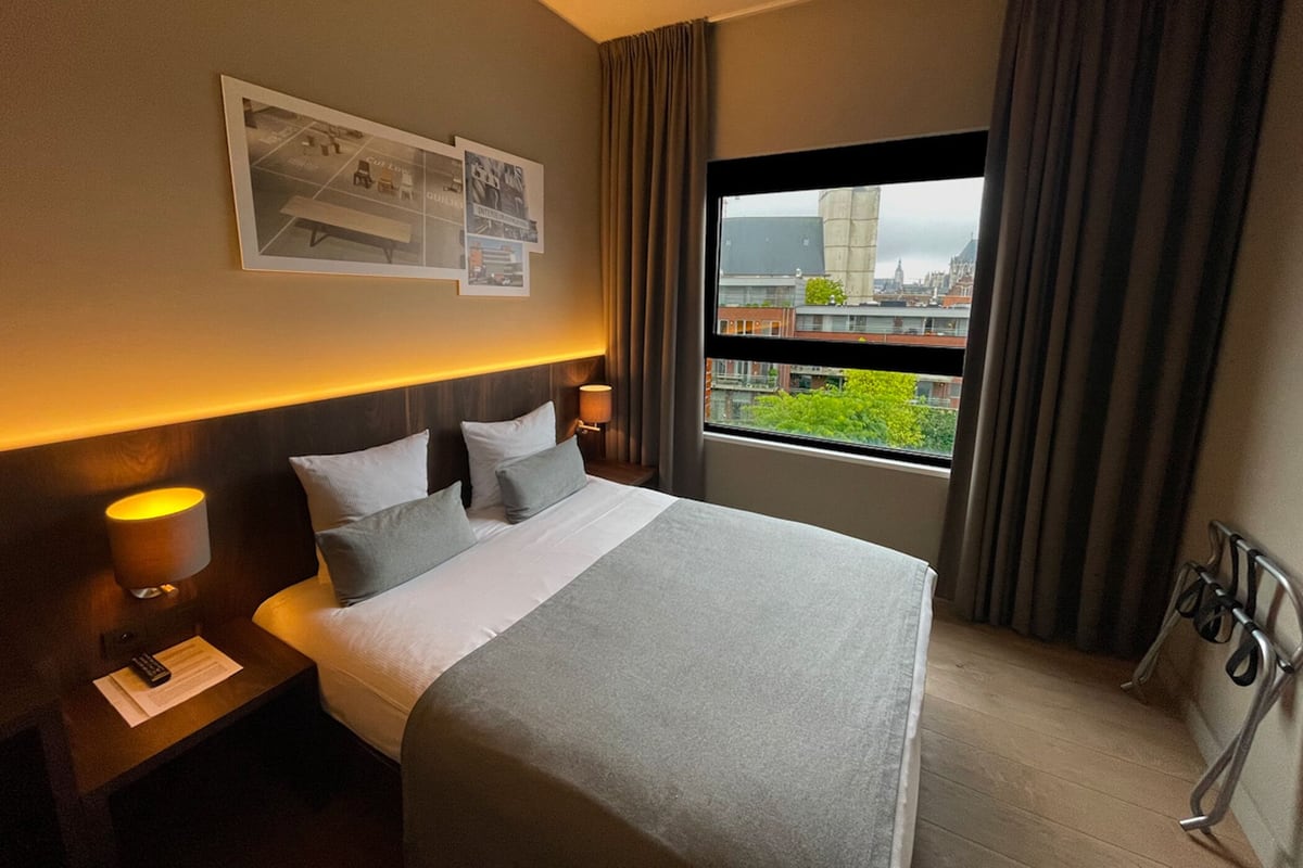 A hotel room with a bed, nightstands, a lamp, a window with curtains, and a city view.