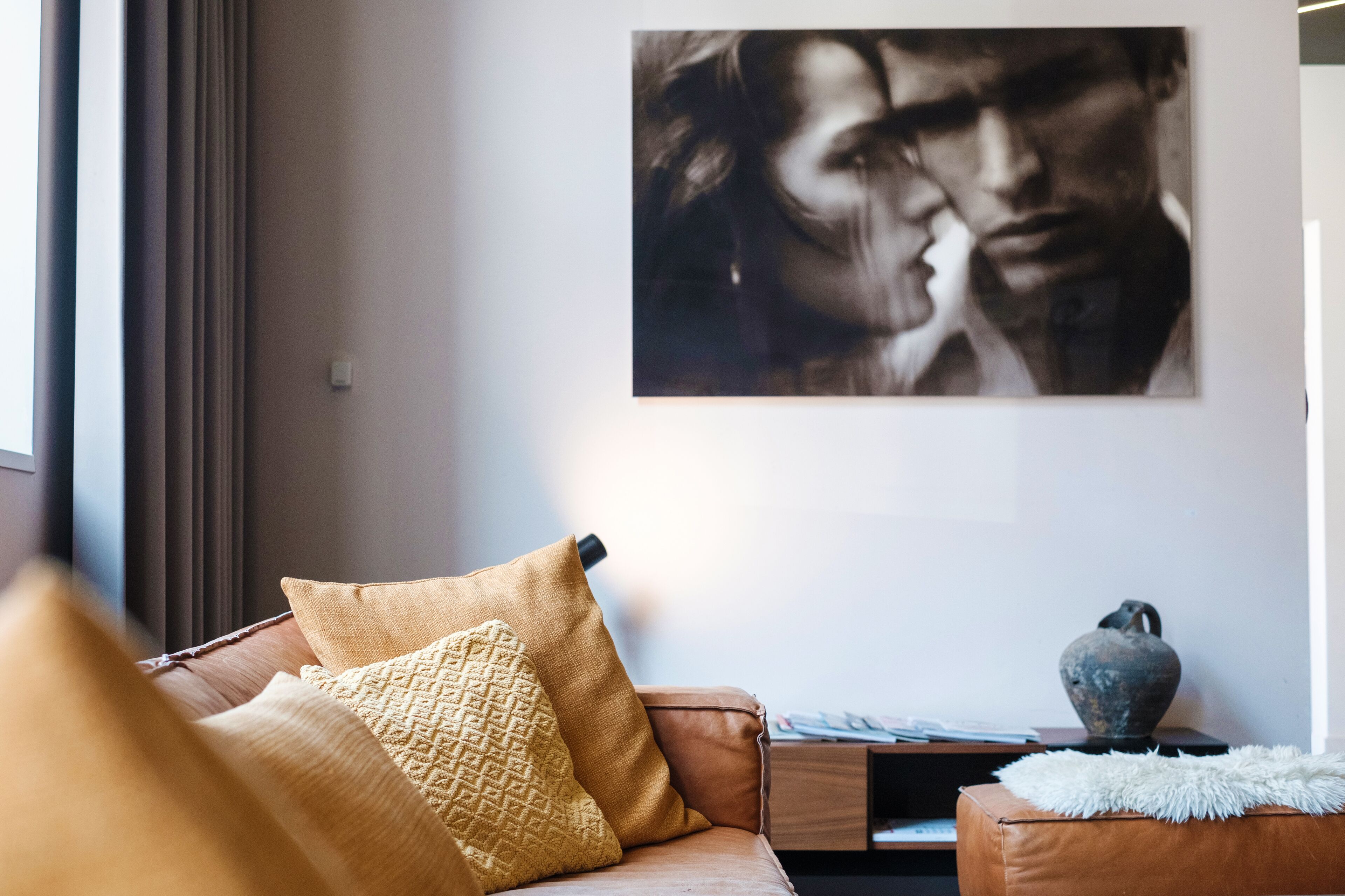 Een woonkamer met een bruine leren bank, een geel gestructureerd kussen, een witte zachte plaid en een vaas op een bijzettafeltje.