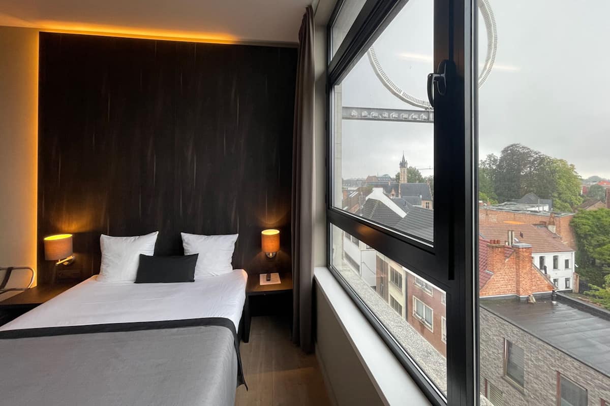 Ein Hotelzimmer mit einem großen Bett, zwei Nachttischen, einem Fenster und Blick auf Gebäude und eine Brücke.