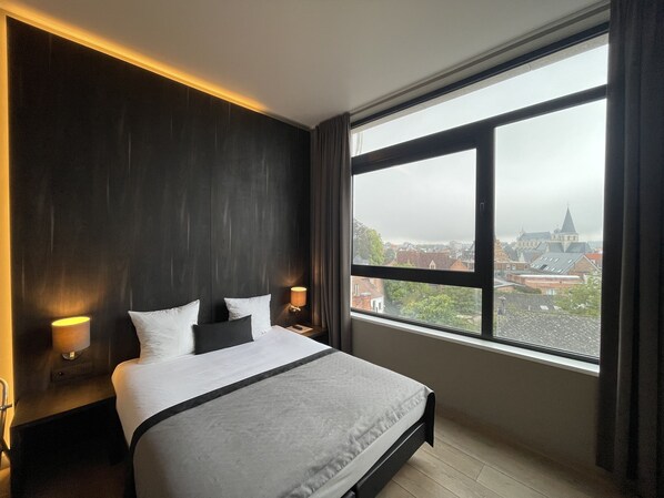 A modern hotel room with a large bed, two bedside lamps, and a view of a cityscape through a large window. - Hotel Elisabeth (Mechelen)
