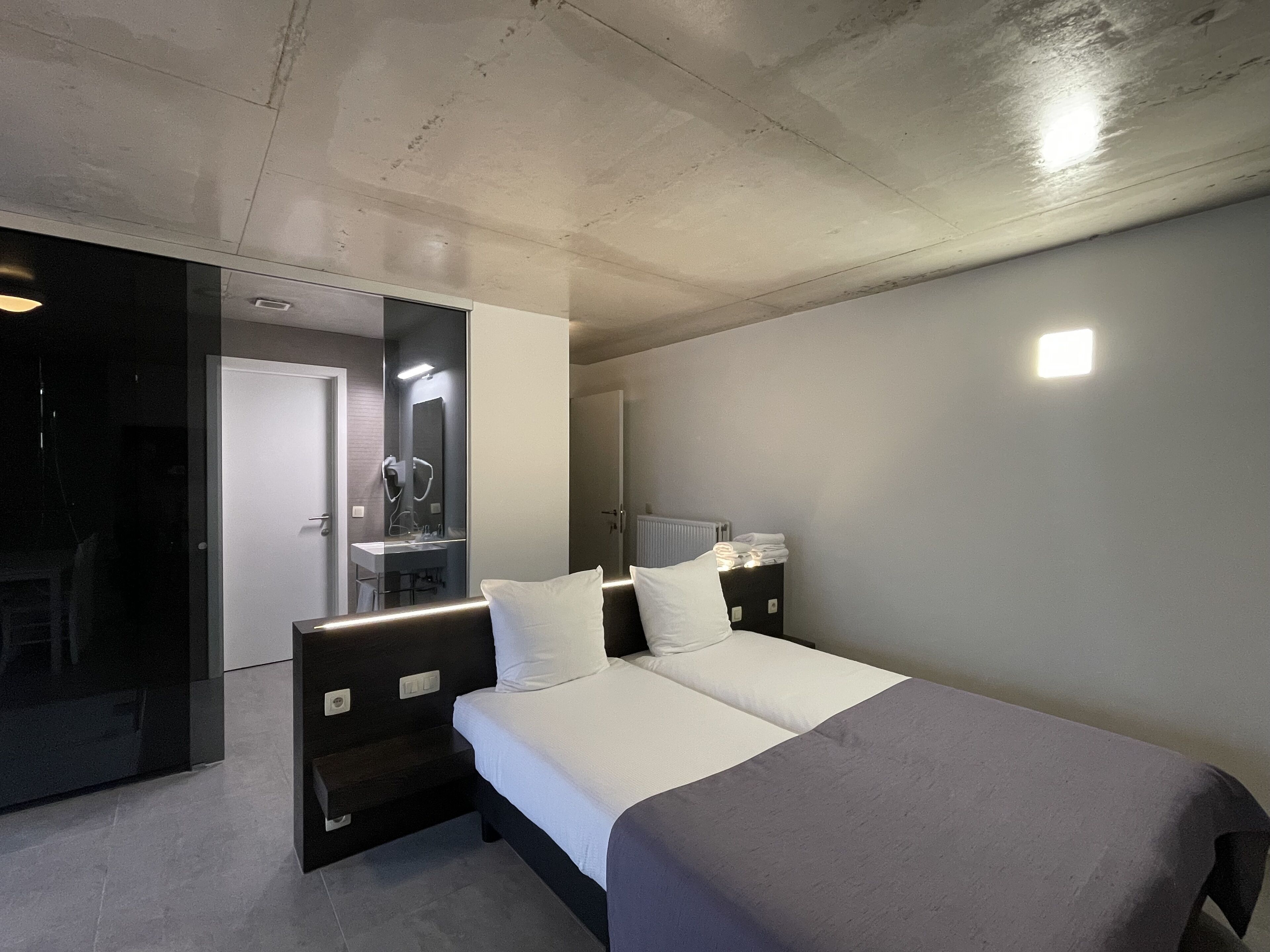 A modern bedroom with a bed, bedside tables, a mirror, and a bathroom visible through an open door.