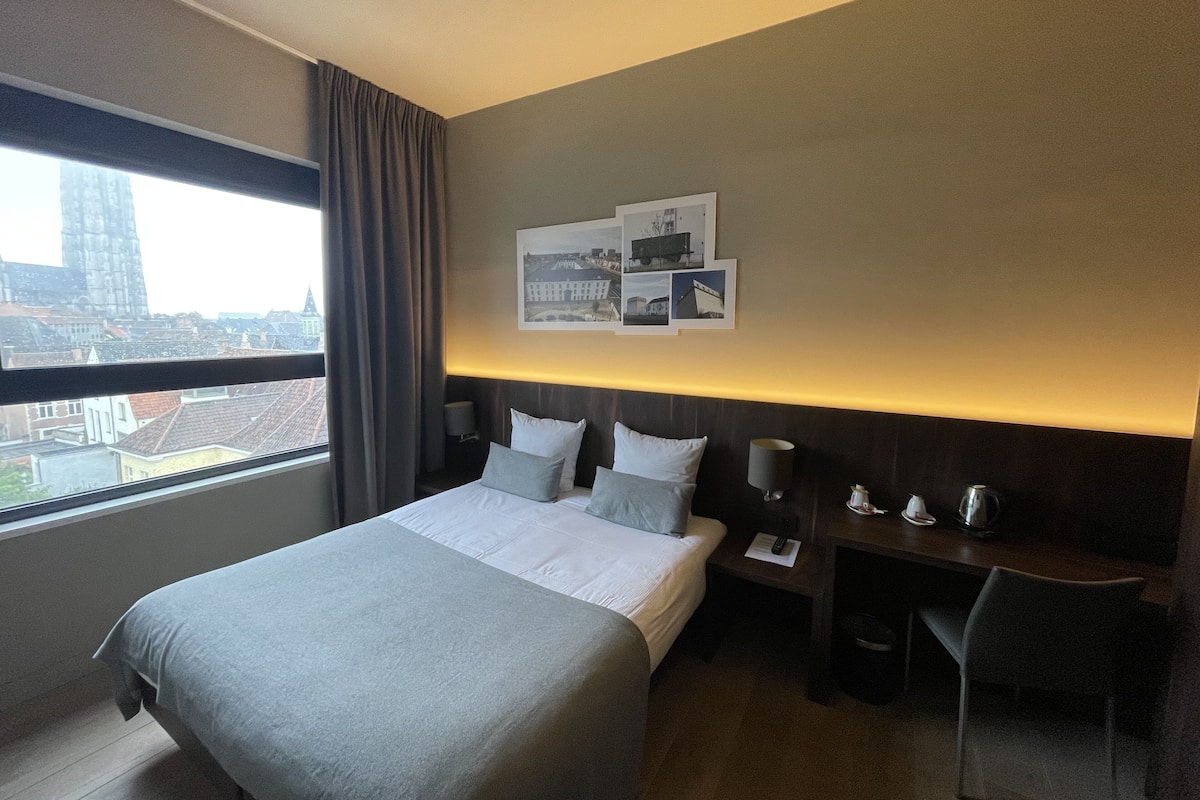 Ein Hotelzimmer mit einem großen Bett, einem Schreibtisch und einem Stuhl. Ein Fenster bietet Blick auf Gebäude.