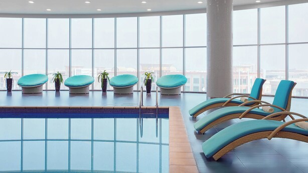 Un área de piscina interior moderna con paredes de vidrio transparente, una piscina con azulejos y varias sillas reclinables con cojines.