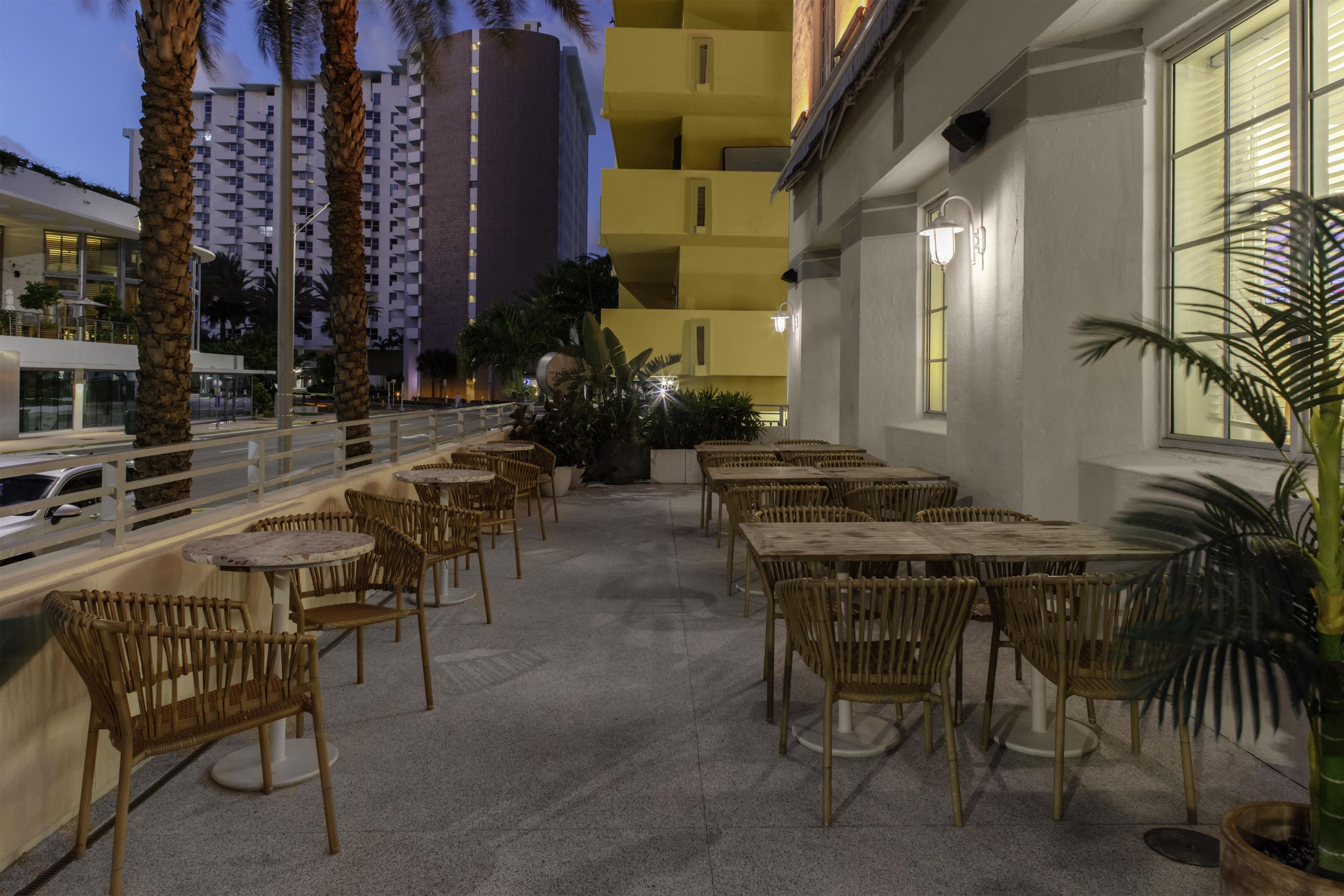 Uma área externa com mesas e cadeiras de vime, corrimão e vista para um horizonte urbano com edifícios e palmeiras.