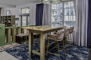 A bar area with a wooden table and bar stools, a green counter with shelves, and a view of the street through large windows. - Hilton Garden Inn Miami South Beach (Miami Beach)