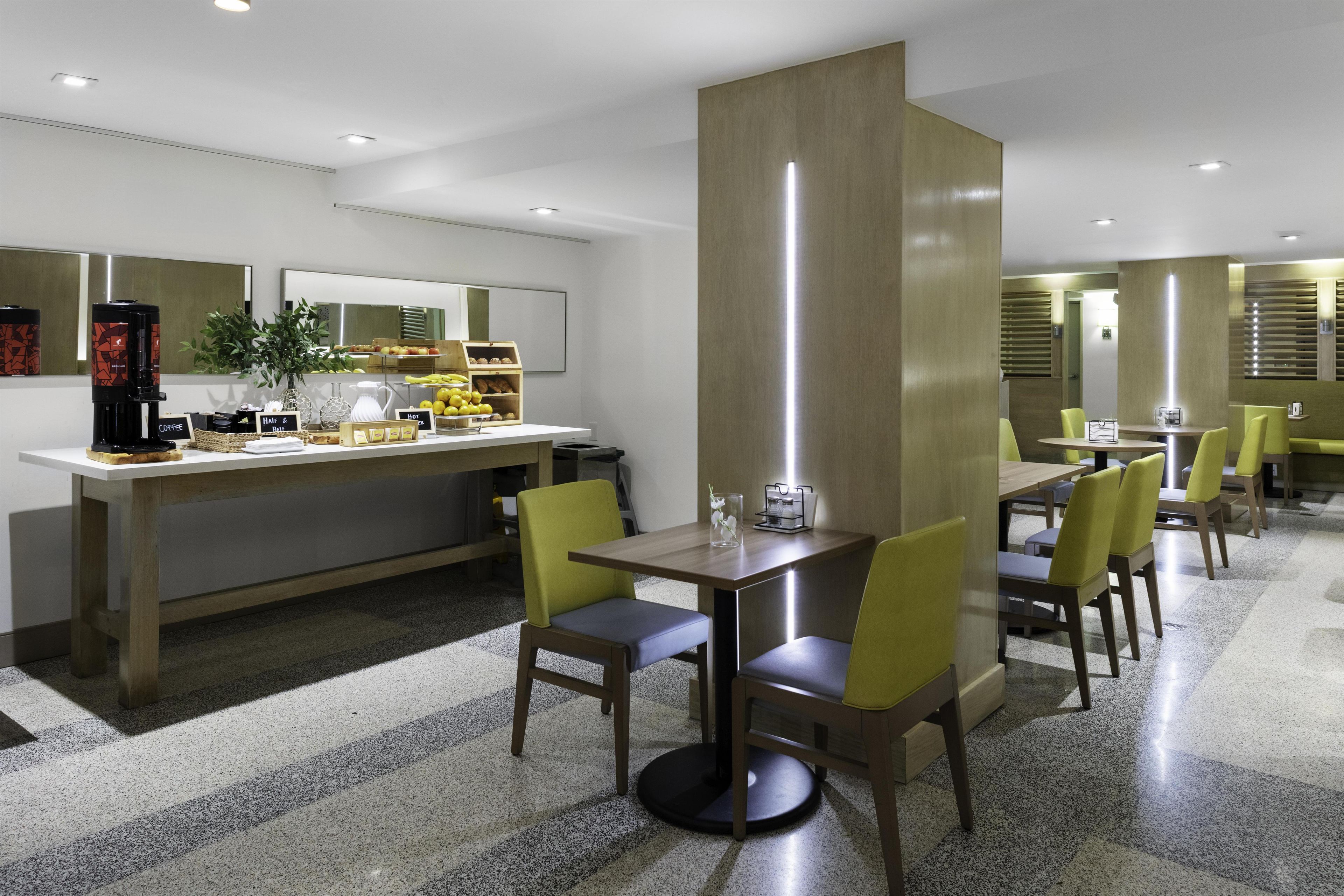 A modern hotel dining area with a coffee station, seating arrangements, and a large mirror.
