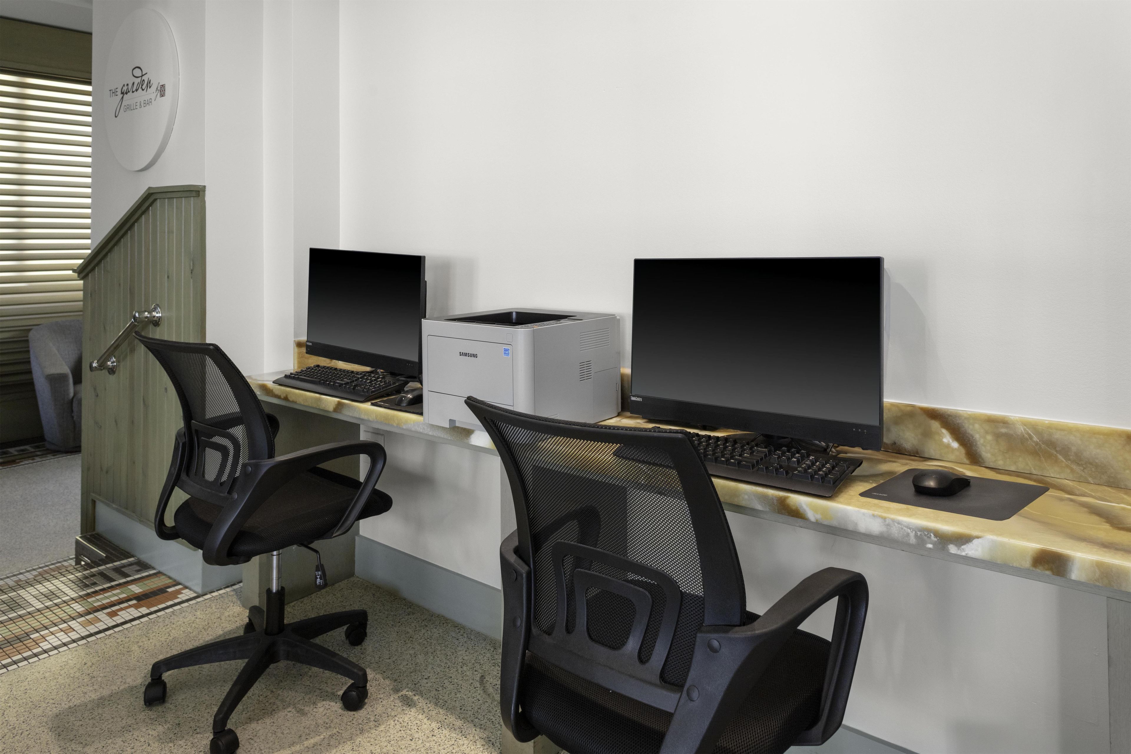 A workspace with two computer desks, each with a monitor, keyboard, and mouse. There are two office chairs. The room has a tiled floor and a wooden door.