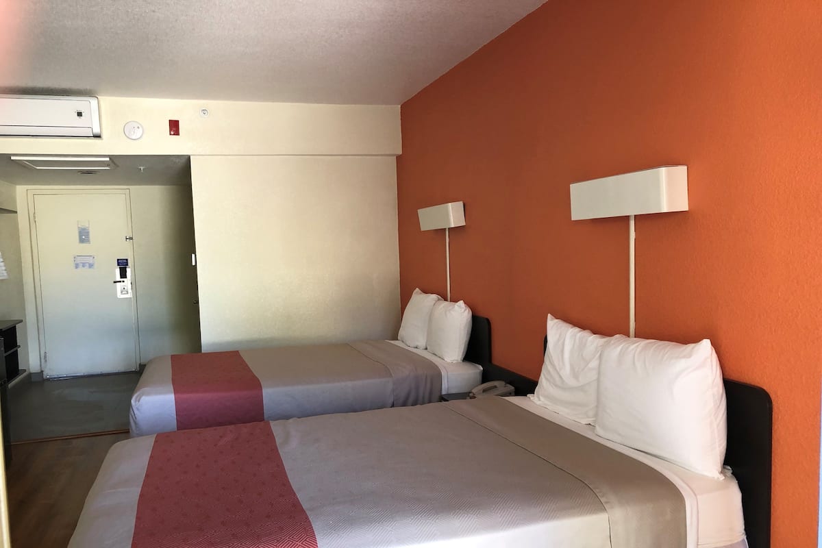 A hotel room with two beds, a door, an air conditioning unit, and a wall-mounted lamp.
