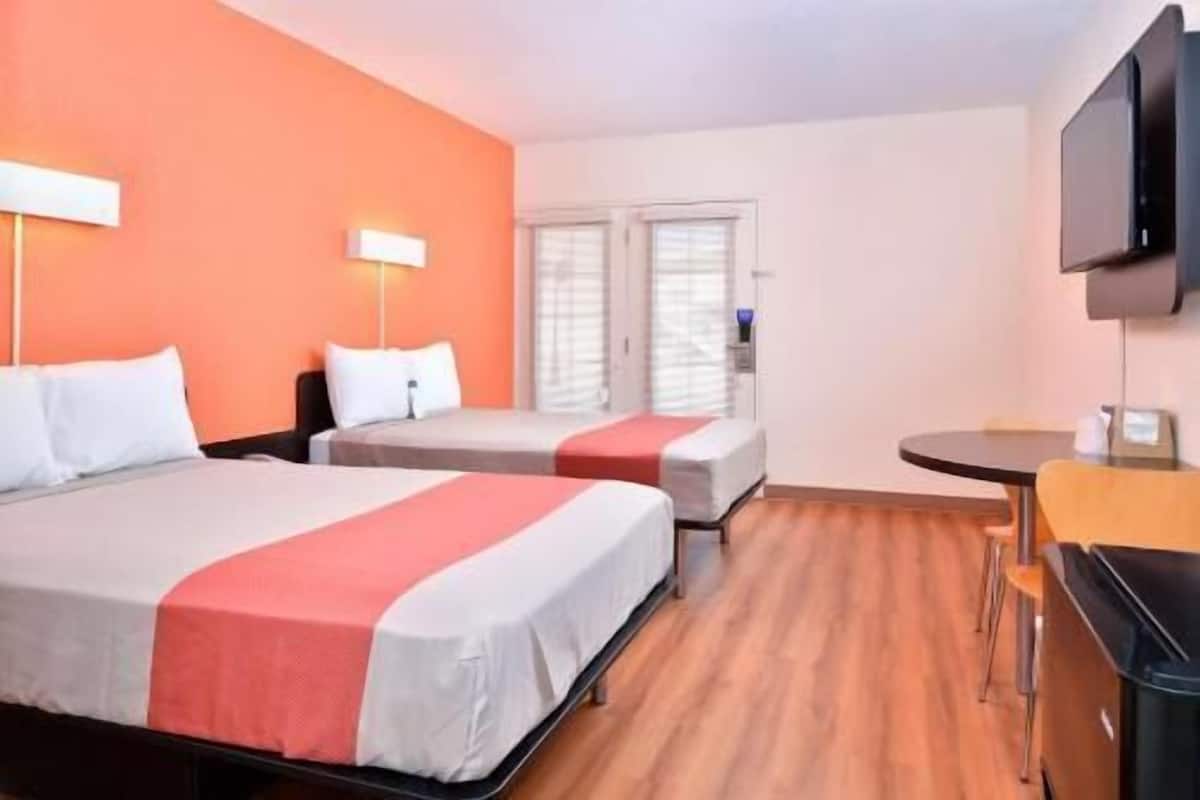 A hotel room with two beds, a small table, a TV, and a window with blinds.