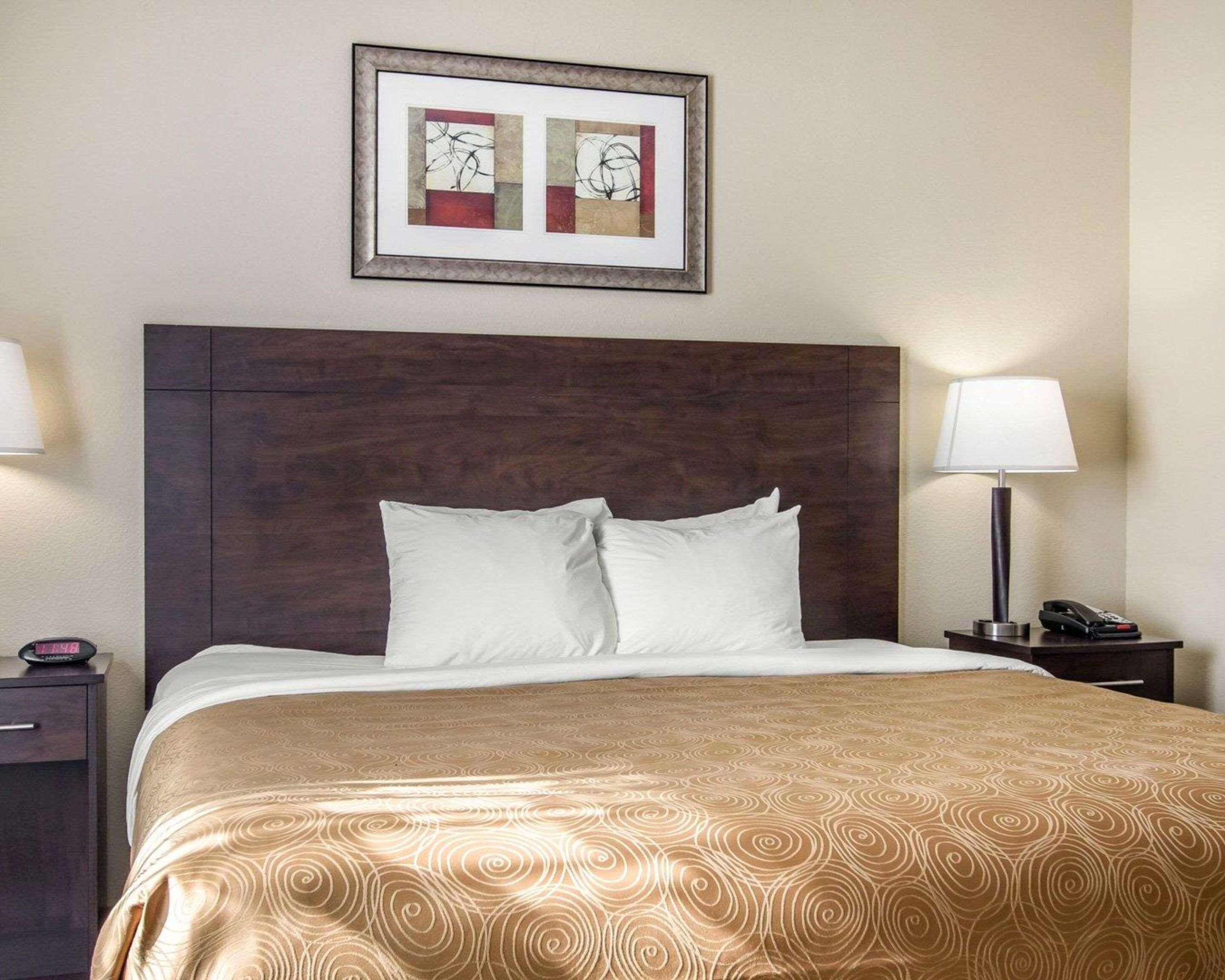A hotel room with a large bed, a dark wooden headboard, two bedside lamps, a framed abstract artwork above the bed, and a nightstand with a phone and a lamp.