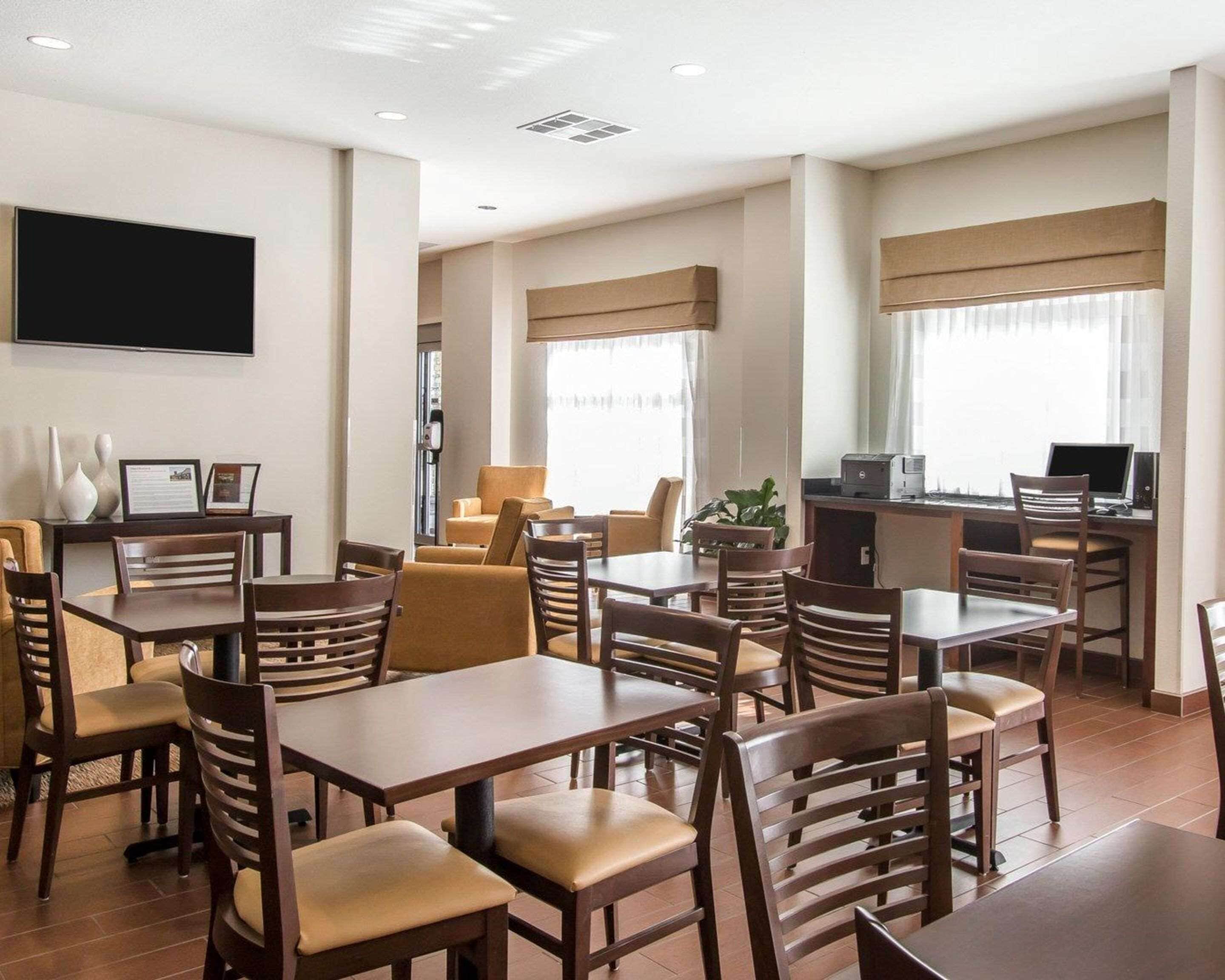 A hotel dining area with multiple tables and chairs, a flat-screen TV mounted on the wall, and a kitchenette area with a computer and printer.