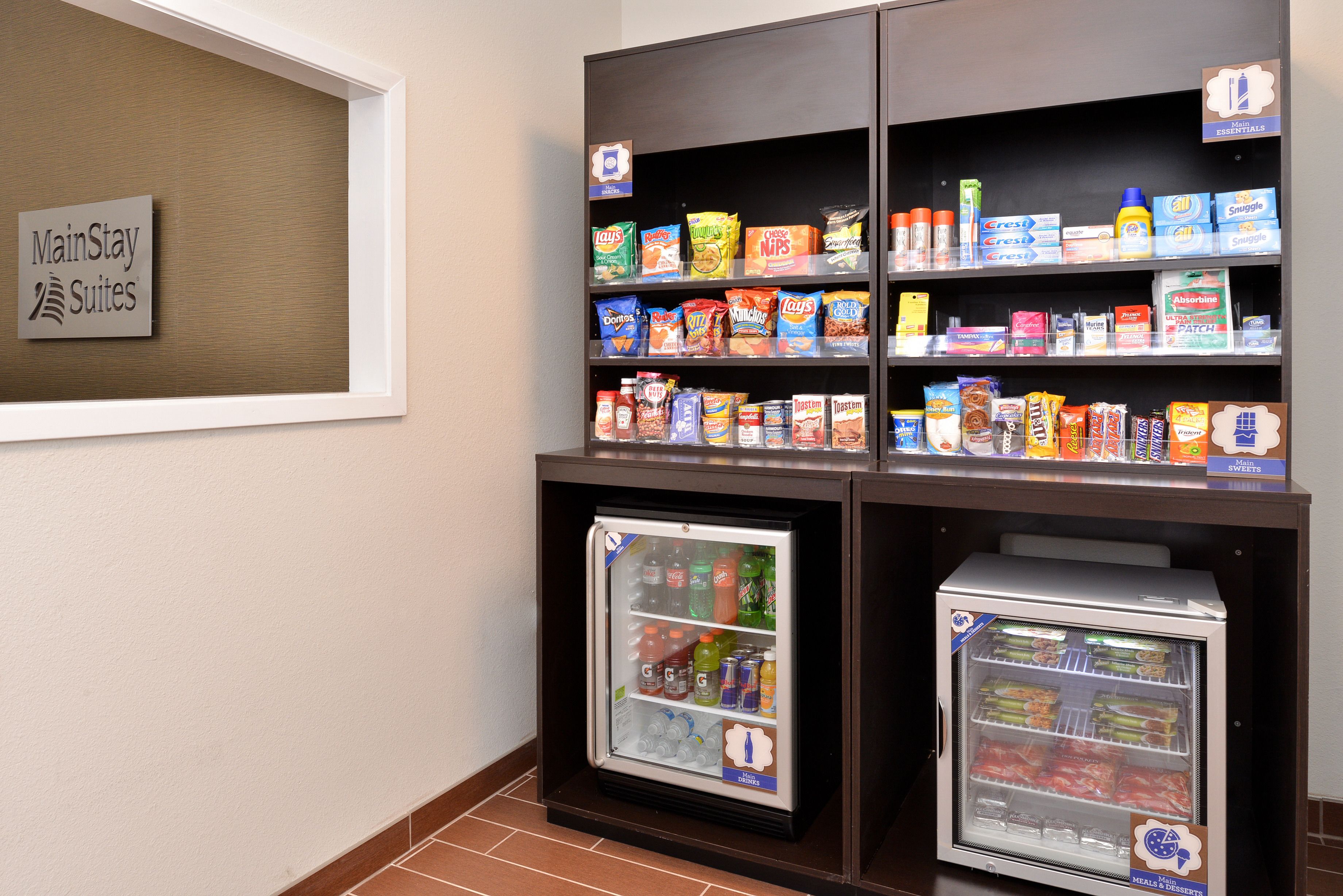 A MainStay Suites mini-fridge stocked with beverages and snacks, a shelf with various food items, and a sign indicating the hotel's name.