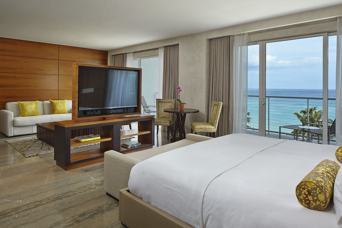A hotel room with a large bed, a TV, a sofa, a small table, and a balcony with a view of the ocean.