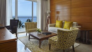 Quarto de hotel moderno com uma grande janela, painel de parede em madeira, sofá com almofadas amarelas e duas cadeiras estampadas.