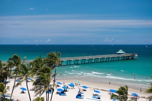 A beach with a pier extending into the ocean, palm trees, and beach umbrellas. - Royal Blues Hotel (Deerfield Beach)