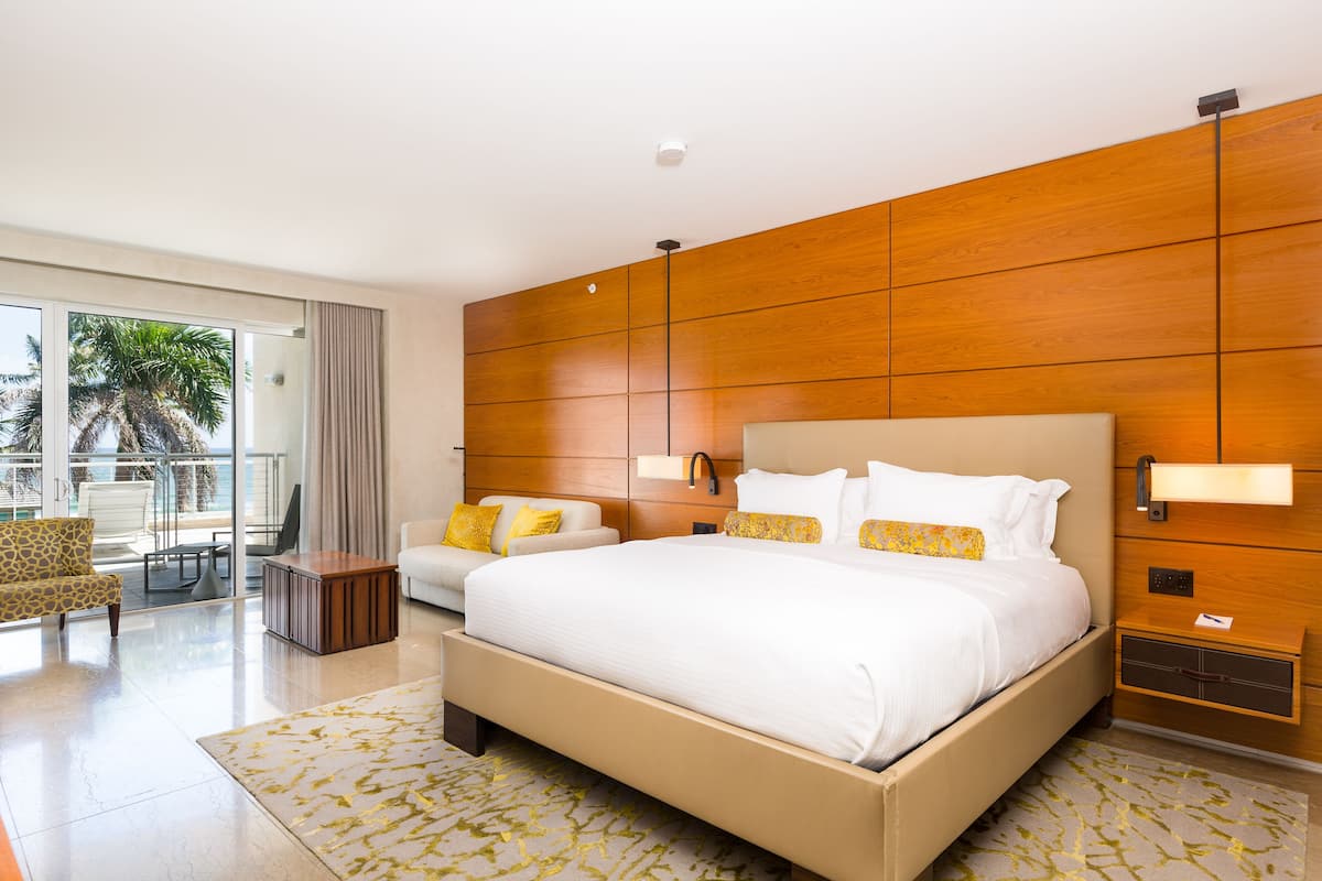 A modern hotel room with a large bed, a sofa, a small table, and a balcony with a view of palm trees.