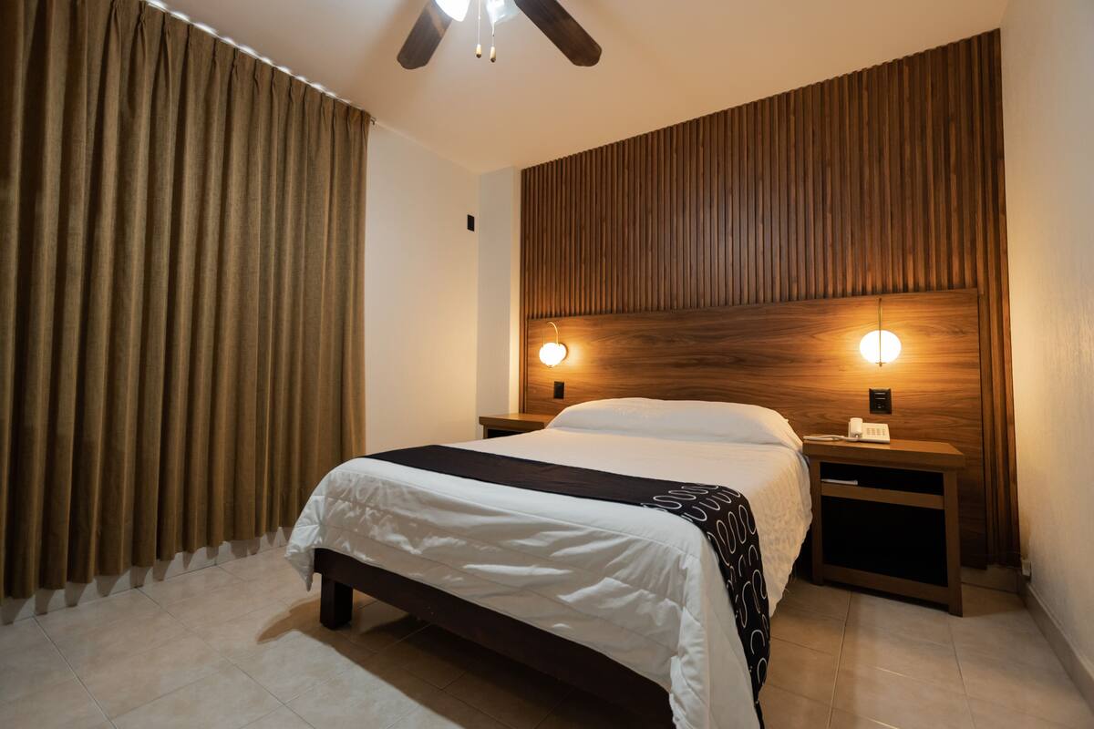 A neatly arranged bedroom with a large bed, wooden headboard, two bedside tables with lamps, and a curtain on the window.