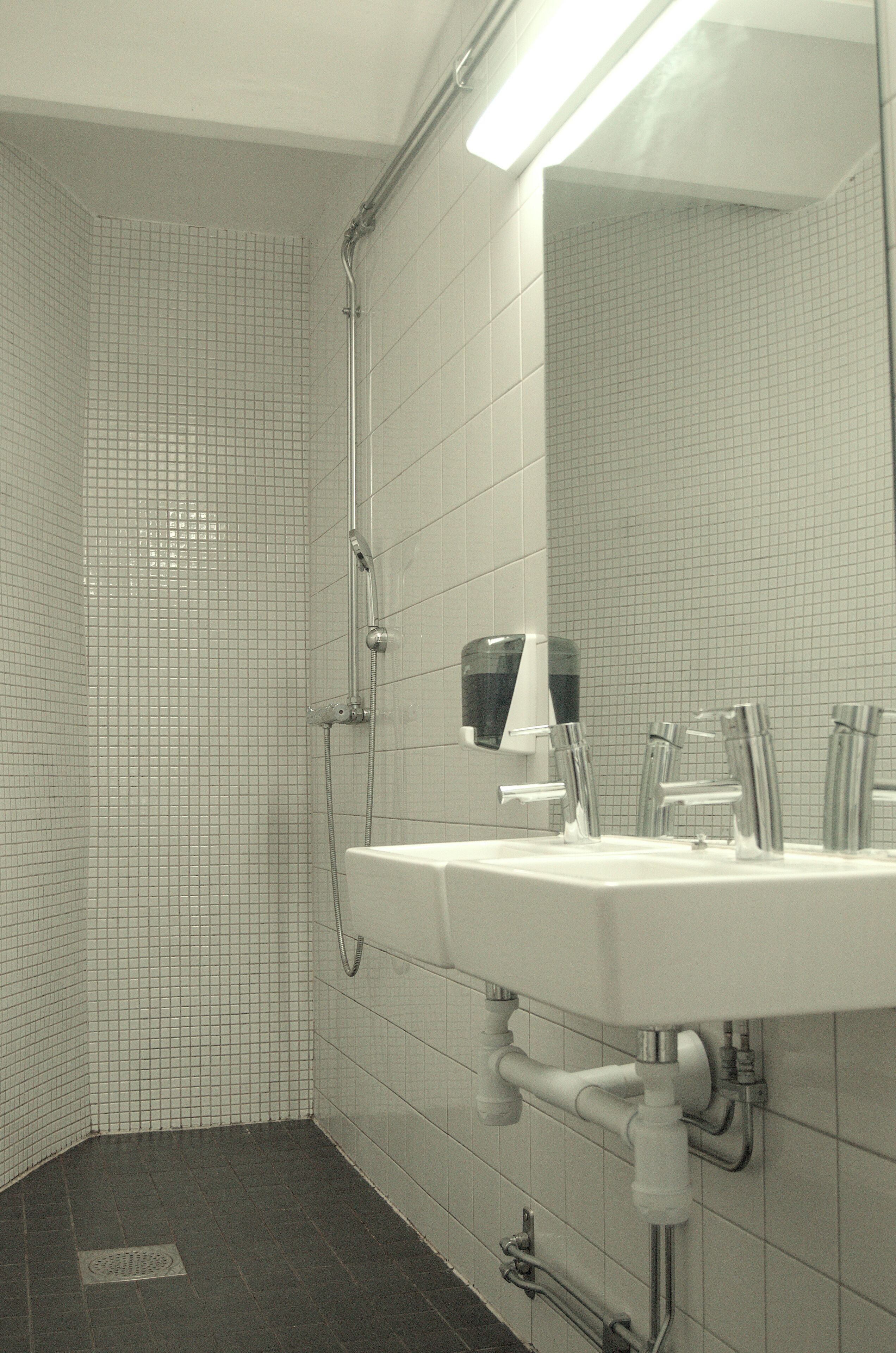 Un baño con paredes de azulejos blancos, un lavamanos blanco con accesorios cromados y una zona de ducha.