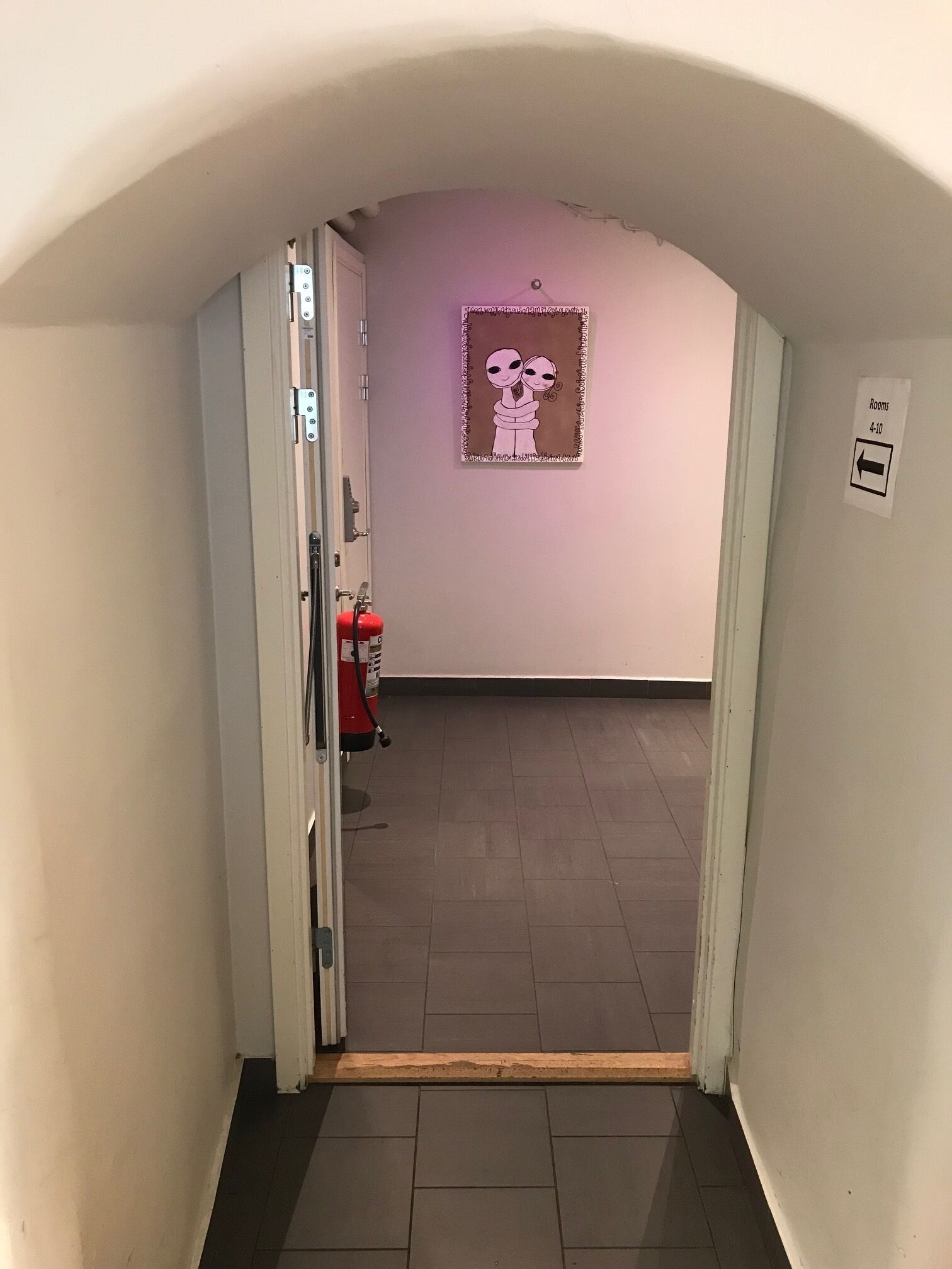 A hallway with a fire extinguisher, a door with a sign indicating rooms 4-10, and a wall-mounted sign with an arrow.
