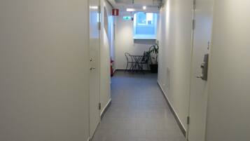 A corridor with white walls, grey tiled flooring, and doors on both sides. There is a small table with chairs and a potted plant in the distance.