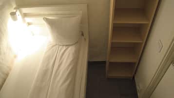 Una cama individual con cabecera blanca, almohada, estantería de madera y suelo de baldosas.
