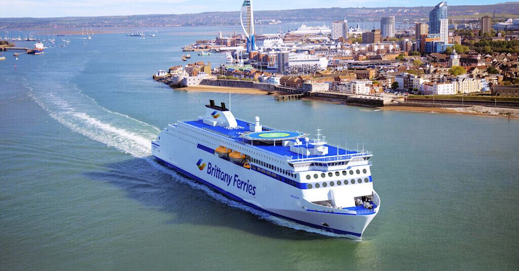 A large ferry named Brittany Ferries traveling through a body of water with a cityscape in the background.