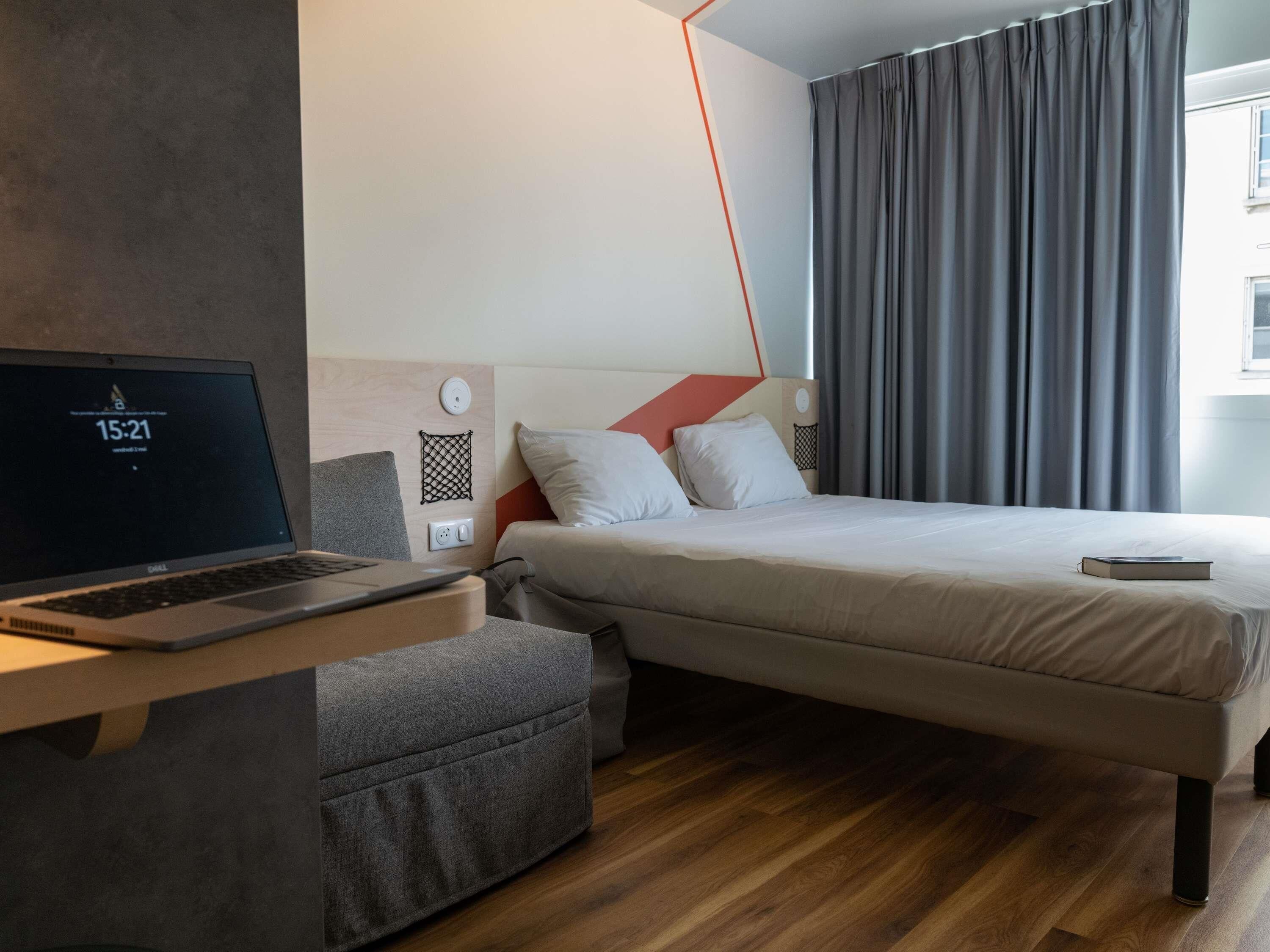 Una habitación de hotel moderna con una cama, un escritorio con una computadora portátil, un enchufe y un pequeño estante en la pared.
