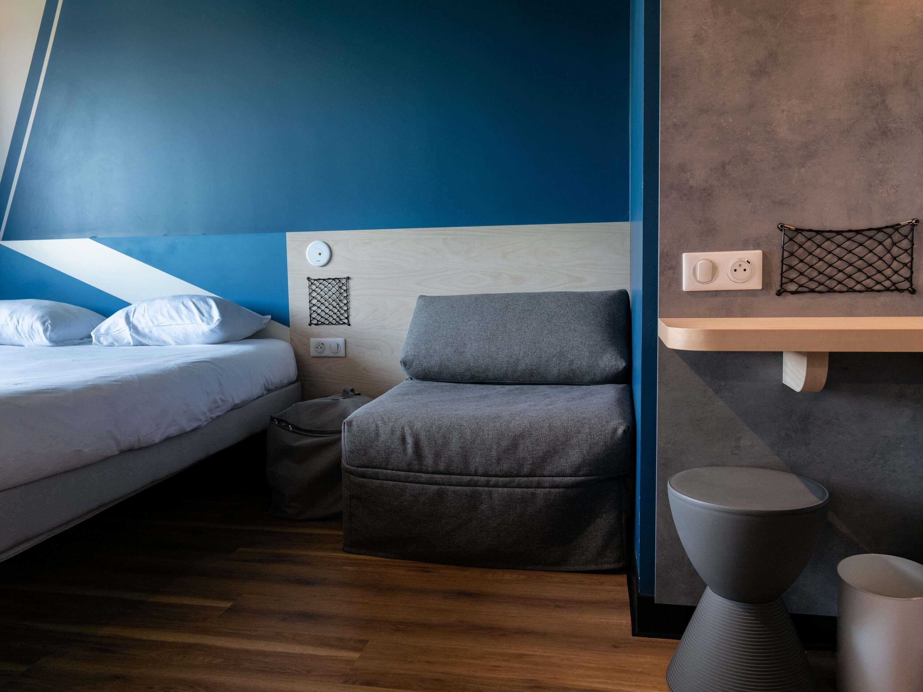 Una habitación moderna con una cama, una silla gris, una mesita pequeña y un estante en la pared.
