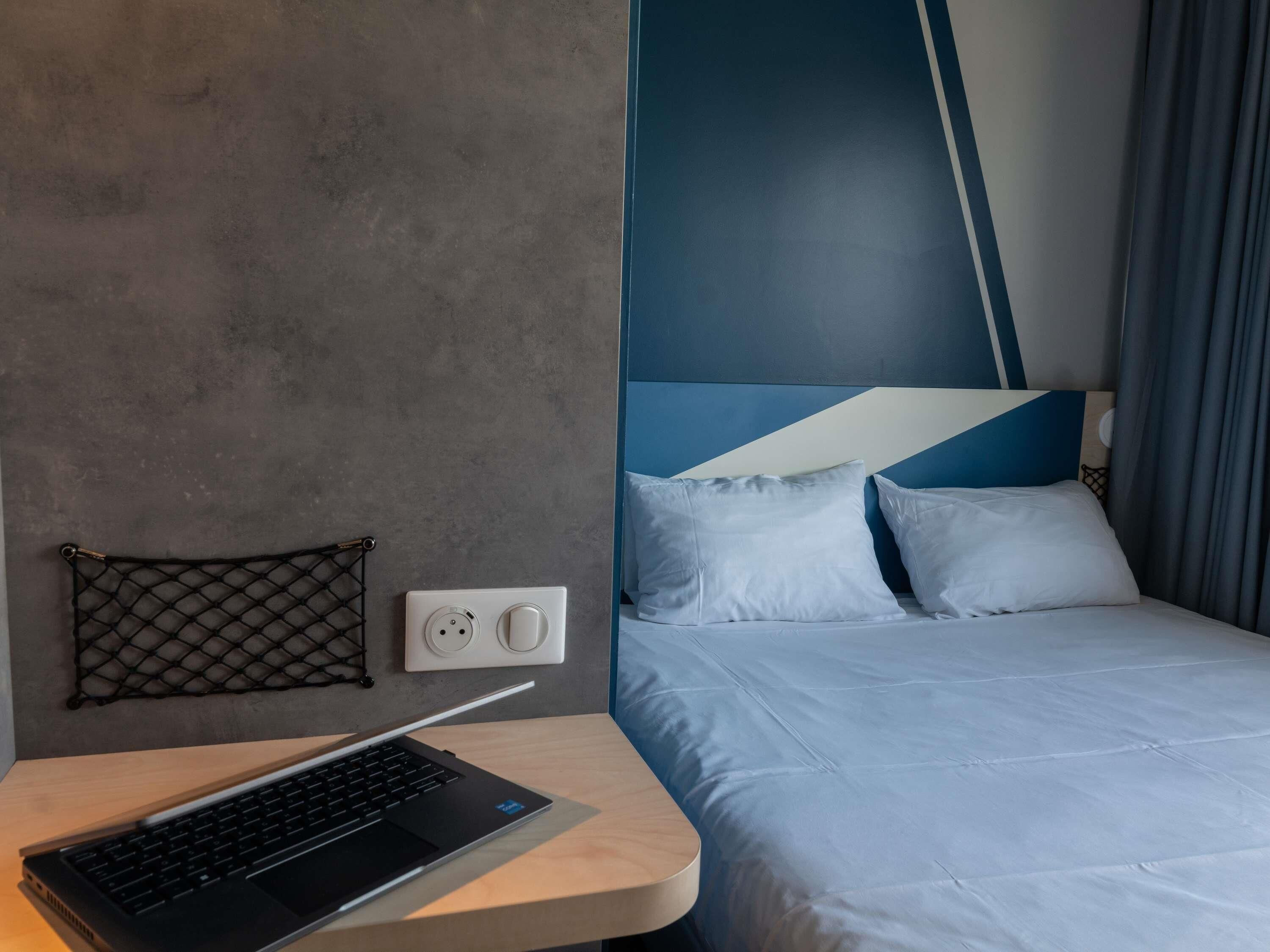 A modern hotel room with a bed, a desk with a laptop, a power outlet, and a wall-mounted light switch.