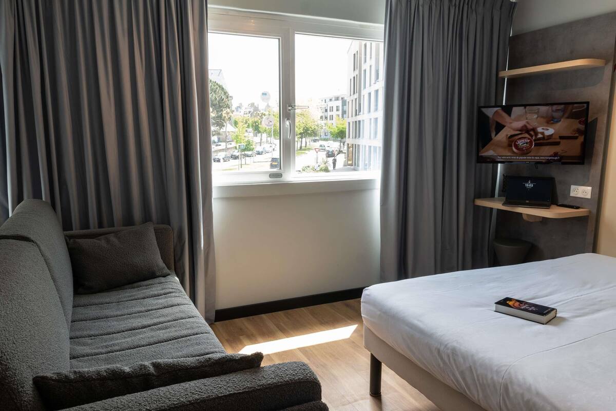 A hotel room with a bed, a sofa, a TV on a shelf, and a window with a view of buildings.
