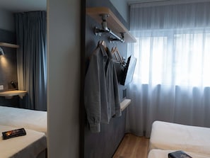 Et hotelværelse med en seng, et fjernsyn ophængt på væggen, et knagered med bøjler, en hylde og et vindue med gardiner.