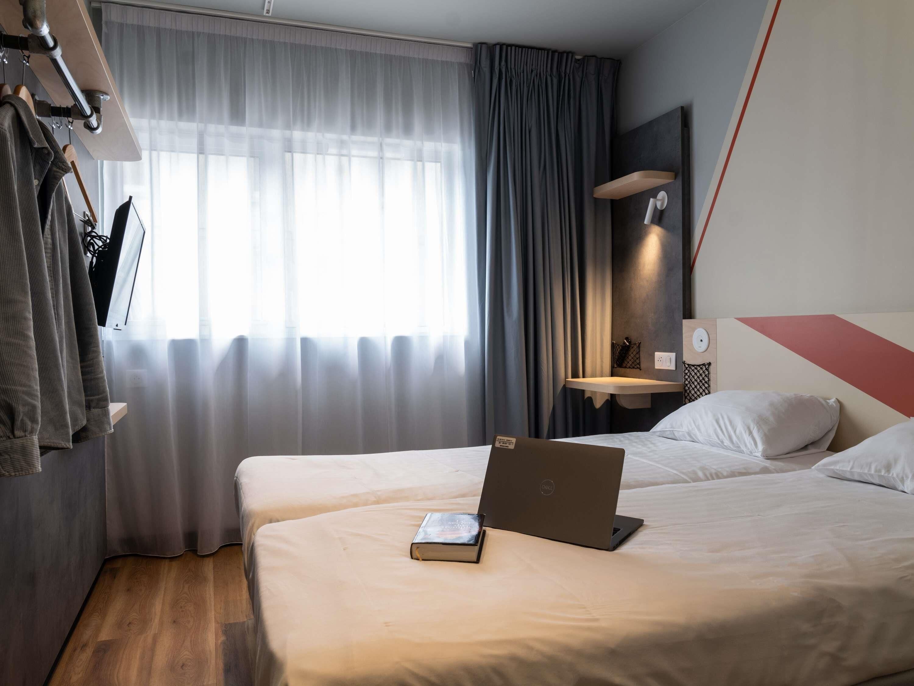 Habitación de hotel con dos camas, una laptop, una mesita de noche, un perchero y una ventana con cortinas.
